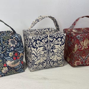May include: Three square fabric bags with top handles, each featuring a distinct botanical and animal print. The left bag is dark blue with birds and strawberries, the centre is cream and navy with rabbits, and the right is red with birds.