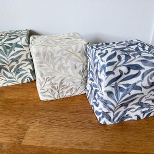 May include: Three square fabric tissue box covers featuring a repeating willow leaf pattern. The left cover is green and cream, the centre cover is beige and white, and the right cover is blue and white. Each box cover has a top opening for tissues.