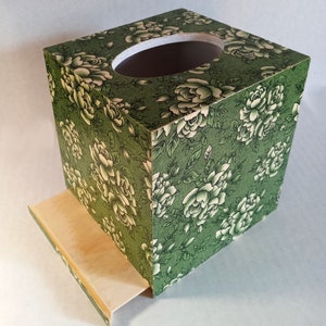 May include: A green tissue box cover with a floral pattern of white roses. The box has a pull-out drawer for easy access to tissues.