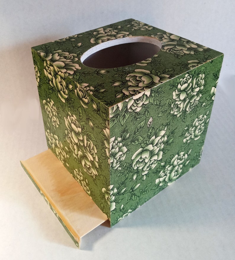 May include: A green tissue box cover with a floral pattern of white roses. The box has a pull-out drawer for easy access to tissues.
