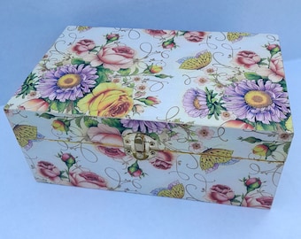 Può includere: Una scatola di legno bianca con un design floreale con rose, margherite e farfalle. La scatola ha una chiusura dorata.