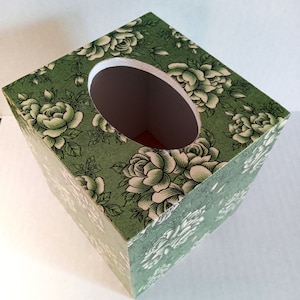 May include: A green tissue box cover with a white floral pattern. The cover has a rectangular shape with a rounded opening on the top.