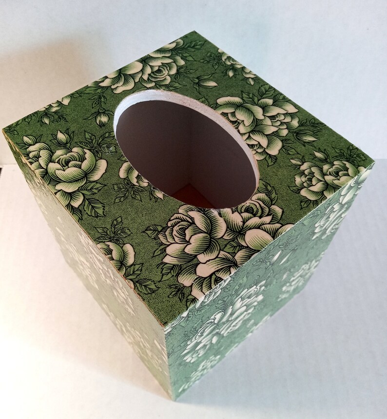May include: A green tissue box cover with a white floral pattern. The cover has a rectangular shape with a rounded opening on the top.