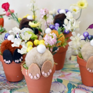 May include: A collection of six small terracotta pots, each decorated with a fluffy bunny tail and felt bunny ears. The pots are filled with colorful spring flowers and greenery.