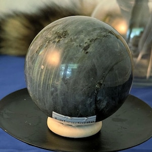 May include: A polished grey and blue labradorite sphere resting on a black metal stand. A small white cylindrical foam piece is attached to the base, featuring the handwritten text 1 inch, indicating the scale of the display stand's central support.