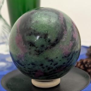 May include: A polished, spherical ruby zoisite stone featuring mottled green, black, and magenta patterns. The sphere rests on a small, light-colored circular stand atop a black metal pedestal. A white cylindrical marker shows a 1 inch measurement.