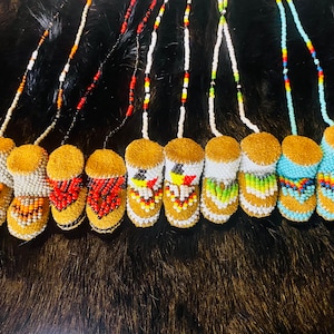 May include: A collection of beaded miniature moccasin pendants. Each pendant features a tan suede top and a beaded body with various colorful patterns. The beadwork includes orange, red, white, and blue beads, strung on beaded cords.