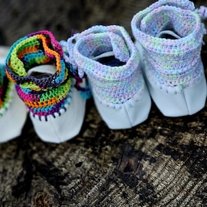 May include: Four pairs of baby booties with white leather uppers and colorful crochet cuffs. The cuffs feature rainbow, pastel, and multi-colored yarn. The booties are arranged on a textured, dark wooden surface.