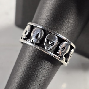 May include: A silver ring featuring a repeating pattern of alien faces. The ring is a band style, with the alien heads evenly spaced around the circumference. The ring is displayed on a dark, textured surface.