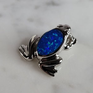 May include: A silver frog charm with a blue opal stone set in the center. The frog is sitting on a white surface.