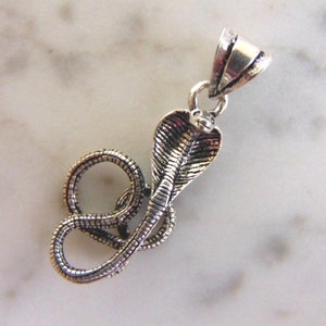 May include: Silver pendant featuring a detailed cobra snake design. The snake is coiled with its head raised and its scales are visible. The pendant has a loop at the top for attaching to a chain.