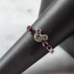 May include: A silver-toned ring featuring a central Mickey Mouse-shaped motif set with three clear stones. The band is accented with small, deep red circular stones embedded along the sides, creating a contrast against the metallic silver finish.