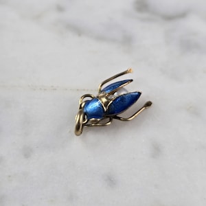 Könnte beinhalten: Ein kleiner, goldfarbener Insektenanhänger mit blau emaillierten Flügeln und einem perlenartigen Akzent. Das Insekt hat zarte Fühler und Beine, vor einem weißen Marmorhintergrund.