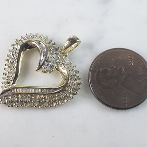 May include: A heart-shaped pendant with a gold-tone frame and a cluster of small, sparkling stones. The pendant is next to a penny for size comparison. The heart's outline is accented with a row of baguette-cut stones.