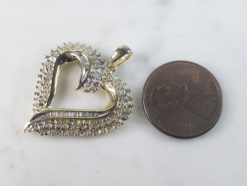 May include: A heart-shaped pendant with a gold-tone frame and a cluster of small, sparkling stones. The pendant is next to a penny for size comparison. The heart's outline is accented with a row of baguette-cut stones.