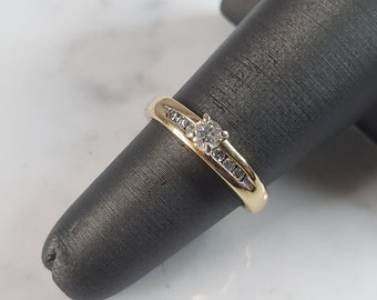 May include: A gold band ring with a single round diamond in the center and smaller diamonds set in a line on either side of the center stone.