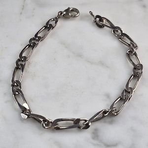 May include: A silver-colored Figaro chain bracelet with an open clasp. The bracelet features a pattern of alternating elongated and oval links. The jewelry is displayed on a white and gray marble surface.