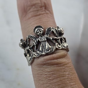 May include: A silver-toned ring featuring a detailed design of angels and floral elements. The ring is worn on a finger, showcasing the intricate craftsmanship and the ring's overall aesthetic. The ring's design includes multiple angels and flowers.