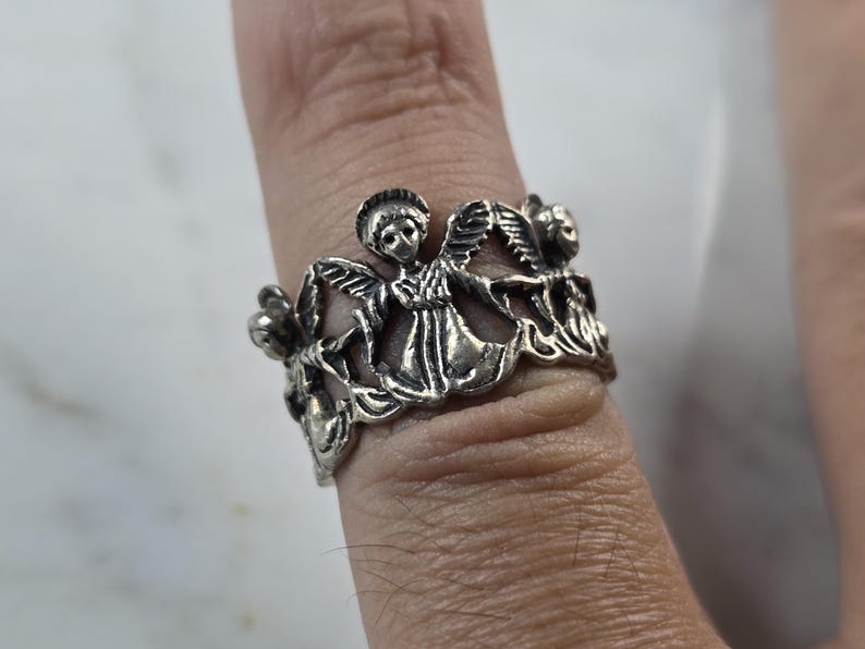 May include: A silver-toned ring featuring a detailed design of angels and floral elements. The ring is worn on a finger, showcasing the intricate craftsmanship and the ring's overall aesthetic. The ring's design includes multiple angels and flowers.