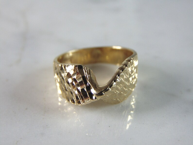 May include: A gold ring with a textured, wavy design. The ring is made of a thick band of metal and has a unique, modern style.