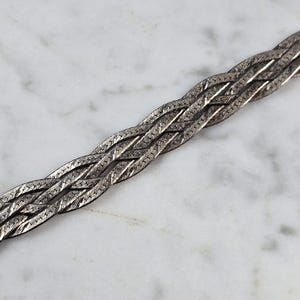 May include: A silver-toned, braided bracelet with a flat, woven design. The bracelet is made of multiple strands woven together, creating a textured surface. The metal has a slightly aged appearance.