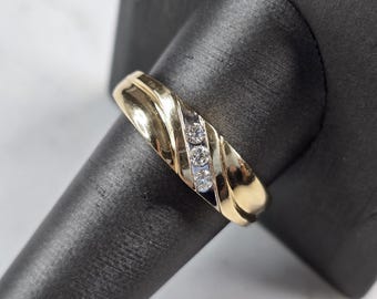Puede incluir: Un anillo dorado con tres pequeños diamantes redondos dispuestos en diagonal. El anillo tiene un diseño texturizado y acanalado en la banda. El anillo se muestra sobre una superficie negra.