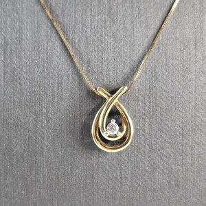 Pode incluir: Um colar com pingente dourado com um design em forma de gota. O pingente apresenta um pequeno diamante redondo no centro. A corrente do colar é delicada e dourada, sobre um fundo cinza.