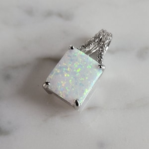 May include: A silver pendant with a white opal gemstone set in a square shape. The pendant has a delicate, twisted design with small clear stones.