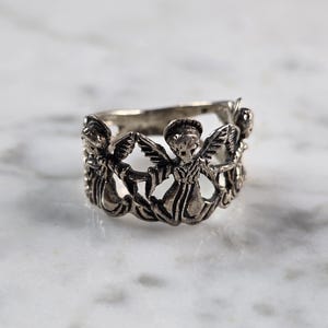 May include: Silver-toned ring featuring a repeating pattern of angel figures with outstretched wings. The ring has a detailed, openwork design, and is set against a marble surface. A piece of jewelry.