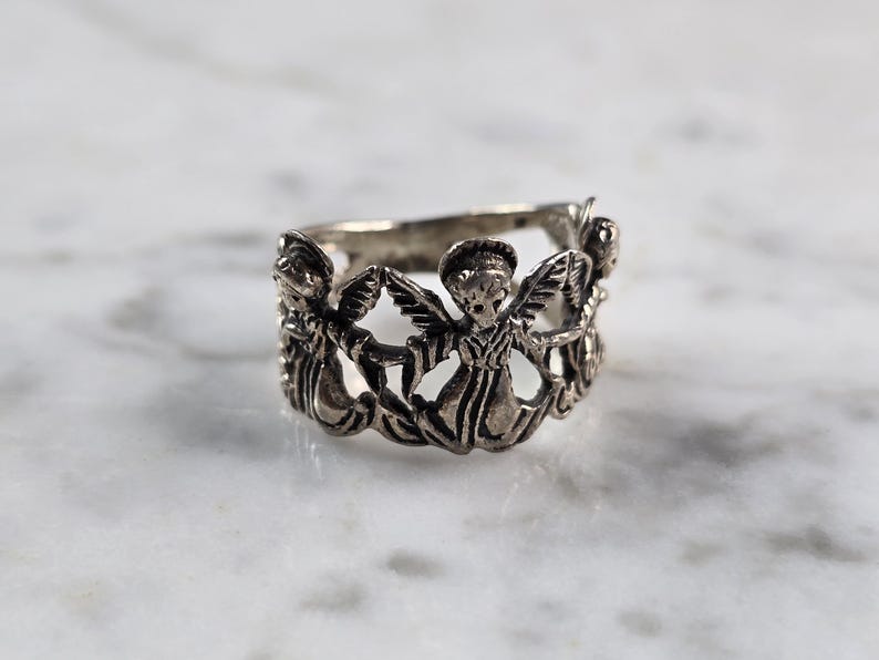 May include: Silver-toned ring featuring a repeating pattern of angel figures with outstretched wings. The ring has a detailed, openwork design, and is set against a marble surface. A piece of jewelry.