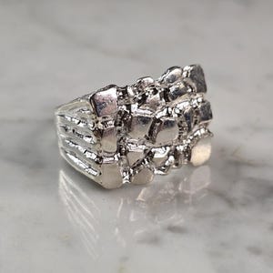 May include: A silver-colored ring with a textured, nugget-like design. The ring has a wide band and a surface composed of irregular, raised shapes. The metal reflects light, creating highlights and shadows. This jewelry item is suitable for everyday wear.