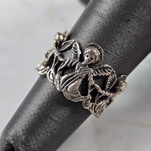 May include: A silver-toned ring featuring a detailed design of cherubs and floral elements. The ring is displayed on a dark gray cylindrical object, showcasing the intricate openwork pattern and the ring's overall aesthetic.