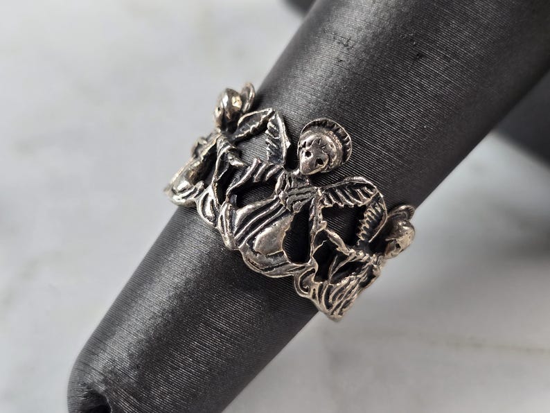 May include: A silver-toned ring featuring a detailed design of cherubs and floral elements. The ring is displayed on a dark gray cylindrical object, showcasing the intricate openwork pattern and the ring's overall aesthetic.