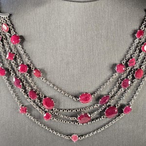 May include: A multi-strand silver necklace with red ruby gemstones. The necklace features a variety of ruby shapes and sizes, including round, oval, and teardrop.