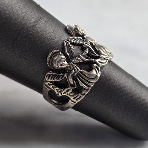 May include: A silver-toned ring featuring a detailed design of figures in a stylized, possibly mythological, motif. The ring is displayed on a dark gray, cylindrical object, showcasing its intricate craftsmanship and vintage aesthetic.