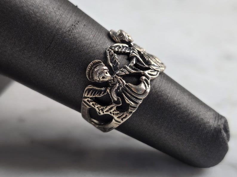 May include: A silver-toned ring featuring a detailed design of figures in a stylized, possibly mythological, motif. The ring is displayed on a dark gray, cylindrical object, showcasing its intricate craftsmanship and vintage aesthetic.