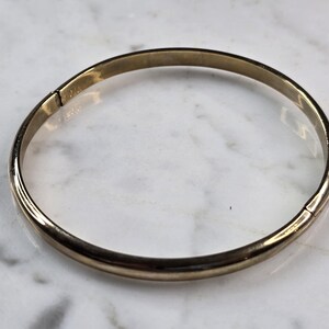 May include: A gold-colored bangle bracelet. The bracelet is a simple, circular design with a hinged clasp. The metal has a slightly worn appearance, suggesting it may be vintage or antique. The bracelet is shown against a white and gray marble surface.