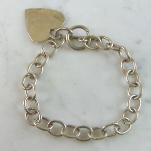 May include: Silver-toned bracelet with a heart-shaped charm. The bracelet features a chain link design with an oval clasp. The heart charm is attached to the chain with a small ring. The bracelet is displayed on a white surface.