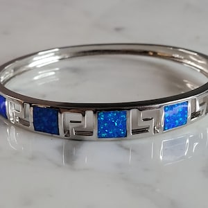 Peut inclure: Un bracelet jonc articulé de couleur argentée orné d'incrustations carrées bleu irisé alternant avec des motifs de grecques ajourés. Le bracelet présente une finition métallique polie et un mécanisme de fermoir sécurisé sur le côté.