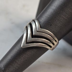 May include: A silver-toned ring with a chevron design. The ring features three stacked chevrons, creating a layered effect. The ring is displayed on a black surface, highlighting its metallic sheen. A piece of jewelry.
