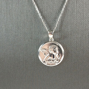 Op de afbeelding: Een zilverkleurige hangerketting. De hanger is een ronde bedel met een gedetailleerd reli&euml;f van een engel. De engel heeft vleugels en rust met zijn hoofd op zijn hand. De ketting is zilver.
