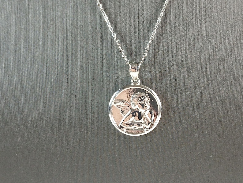 Op de afbeelding: Een zilverkleurige hangerketting. De hanger is een ronde bedel met een gedetailleerd reli&euml;f van een engel. De engel heeft vleugels en rust met zijn hoofd op zijn hand. De ketting is zilver.