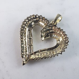 May include: A heart-shaped pendant with a halo of small, round gemstones and a central row of rectangular gemstones. The pendant is made of gold-colored metal and has a loop for a chain.