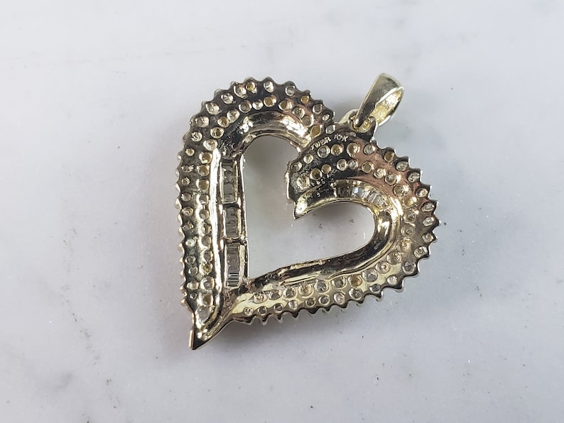 May include: A heart-shaped pendant with a halo of small, round gemstones and a central row of rectangular gemstones. The pendant is made of gold-colored metal and has a loop for a chain.