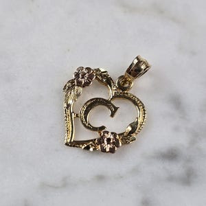 May include: Gold-tone heart pendant with the letter "G" in the center. The pendant features floral and leaf accents in a contrasting rose gold color. The pendant is designed for jewelry.