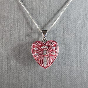 May include: A silver chain necklace with a red heart-shaped locket. The locket has a silver butterfly design with clear rhinestones.