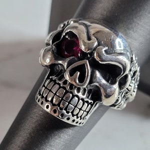 May include: A silver skull ring with a red gemstone eye. The skull is detailed with a heart shape on the forehead and a row of teeth.
