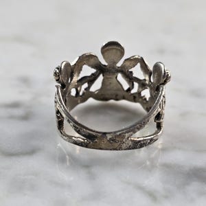 May include: A silver-toned ring with an ornate design. The ring features a central figure with outstretched arms, flanked by decorative elements. The ring's band has a double-layered design. The ring is set against a marble surface.