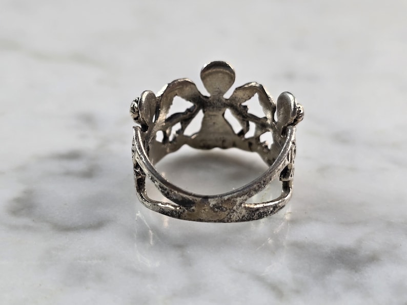 May include: A silver-toned ring with an ornate design. The ring features a central figure with outstretched arms, flanked by decorative elements. The ring's band has a double-layered design. The ring is set against a marble surface.