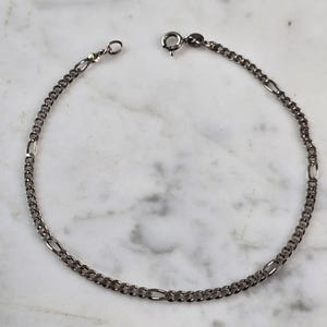 May include: A silver-toned chain bracelet with a lobster clasp closure. The bracelet features a combination of small, rounded links and elongated, flattened links, creating a textured pattern. The bracelet is displayed on a white and gray marble surface.